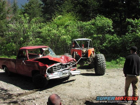 Red pickup truck with smashed front end being towed by an orange off-road buggy. Person watches