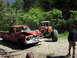 1993 Ford Bronco - A severely damaged red pickup truck is being towed by a large off-road vehicle on a dirt road.