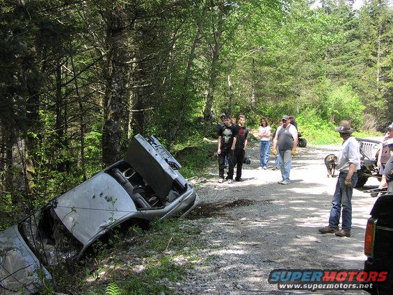 Car flipped on its side in a wooded area with people standing around