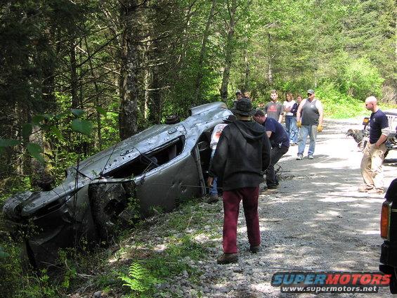 Overturned silver SUV on its side in a wooded area with several people around it