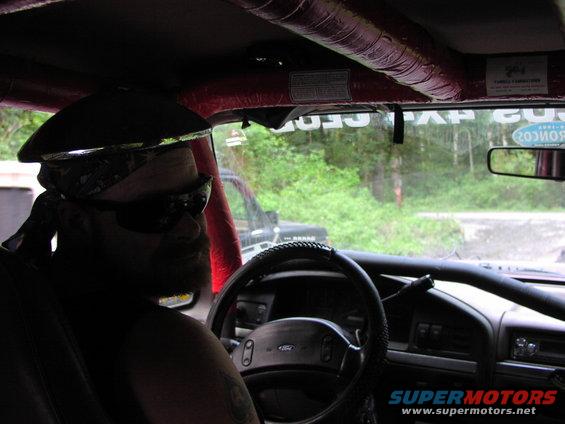 Driver wearing bandana and sunglasses inside a 1993 Ford Bronco with roll cage. Another Bronco is visible outside