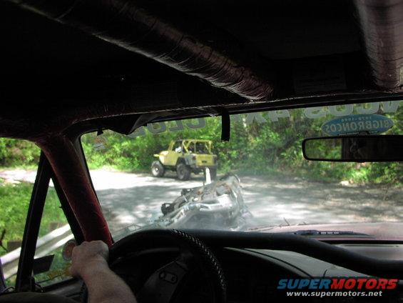 Interior view of a red vehicle looking at an overturned vehicle and a yellow Jeep on a dirt road