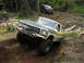 1993 Ford Bronco - Two Ford Broncos off-roading on a dirt path with trees and logs surrounding them.