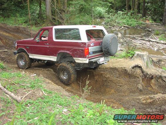 Maroon Ford Bronco with white top driving on a muddy trail, going over a ditch. Spare tire mounted on the back