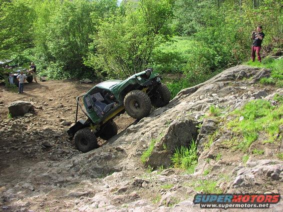 Green off-road vehicle climbing a steep, rocky incline with trees in the background. Another vehicle is visible to the left