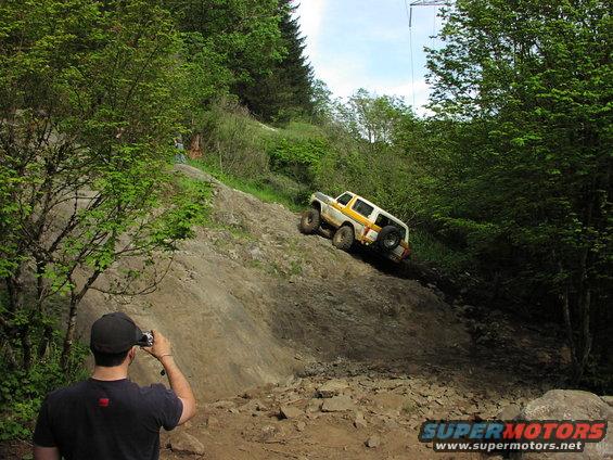 White and yellow Ford Bronco climbing a rocky incline, with a person photographing it from below