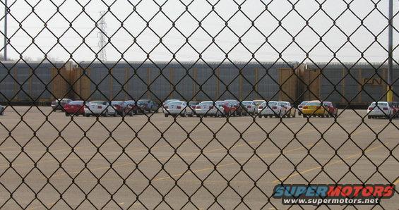 Cars parked in a lot with a train in the background, viewed through a chain-link fence