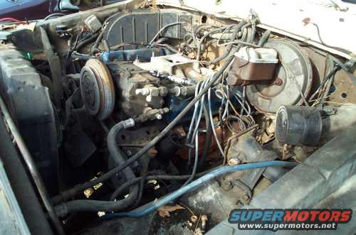 Engine bay with V8 engine, radiator, and brake booster. Visible hoses and wiring. Some rust and dirt