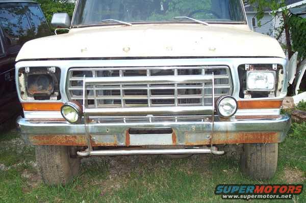 Front view of a white Ford truck with a chrome grille and rusty bumper. Headlights are missing
