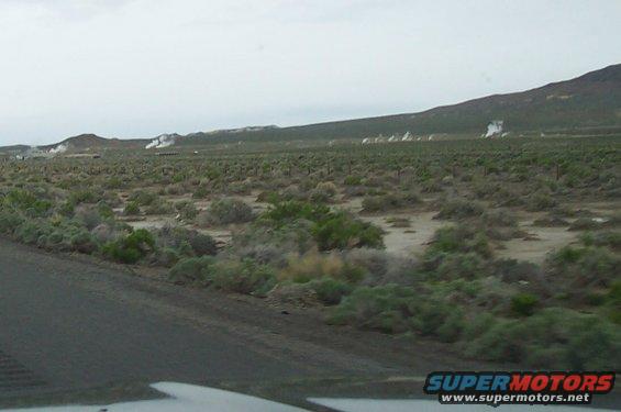 Steam rising from geothermal vents in a vast, arid landscape with low brush and distant mountains