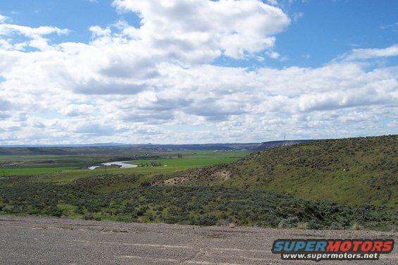 Gravel road in foreground, rolling green hills, river, and distant mountains under a cloudy blue sky