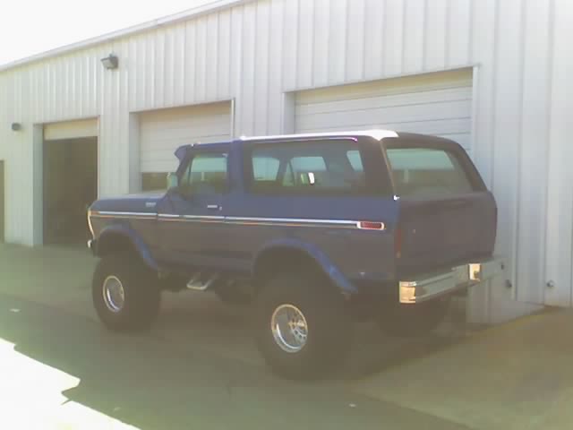 Blue 1978 Ford Bronco with chrome trim and aftermarket wheels parked in front of a metal building