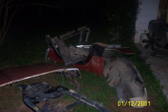 Pile of disassembled car parts, including a red fender and black frame pieces, on grass