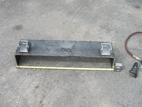 Metal bracket with two mounting tabs, a yellow tape measure, and an air hose fitting on asphalt