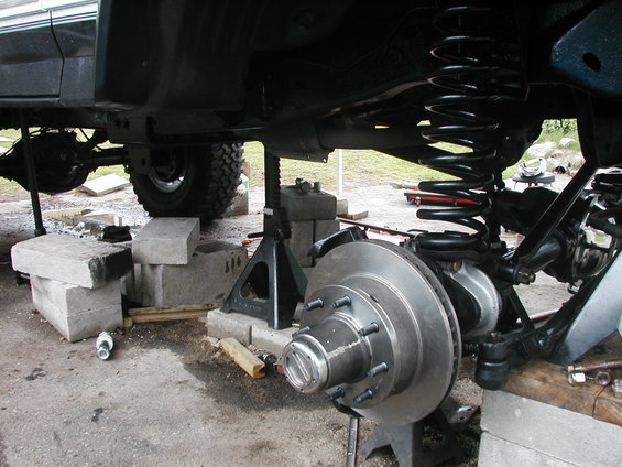 Vehicle undercarriage with suspension and brake components visible, supported by jack stands and blocks