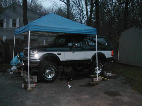 Dark green and white Ford Bronco lifted on blocks under a blue canopy