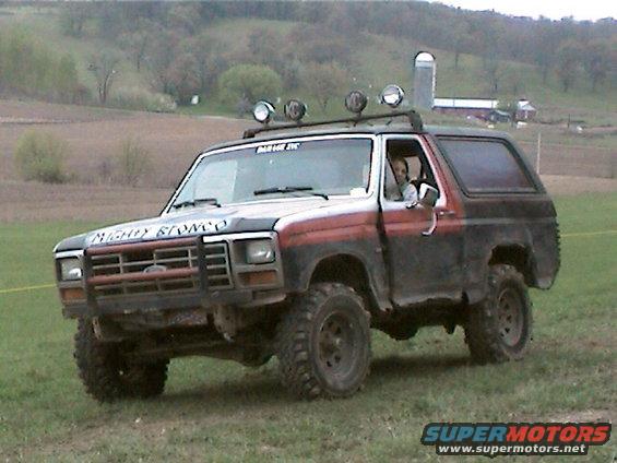 Two-tone red and black Ford Bronco with off-road tires and roof-mounted lights driving on a grassy hill