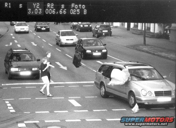 Traffic camera shot of people crossing street, cars stopped, one car has a large white bag sticking out of the window
