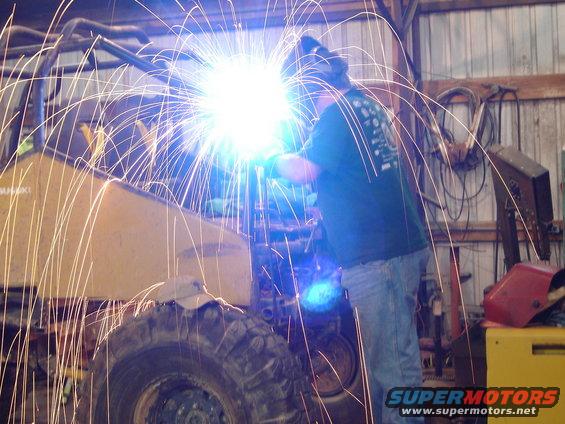 Welding sparks fly around a yellow vehicle frame and large tire