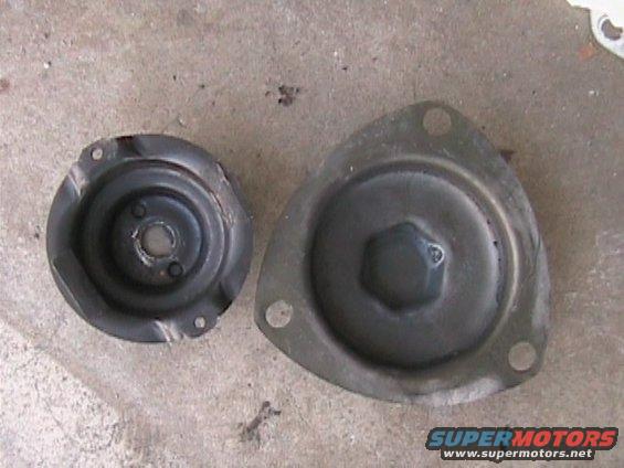 Two black metal car suspension parts on a concrete surface. One is circular with a damaged rim, the other is triangular with a hexagonal center