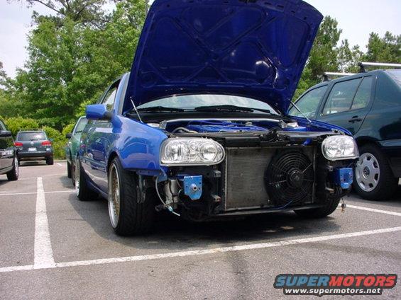Blue car front view, hood open, engine exposed. Other cars parked nearby