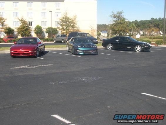 Three sports cars parked in a lot: a red Ford Probe, a teal modified car with hood open, and a black car with 'GTX' badge