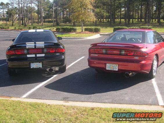 Two Ford Probes parked in a lot, one black with white stripes, one red. Both have racing stripes
