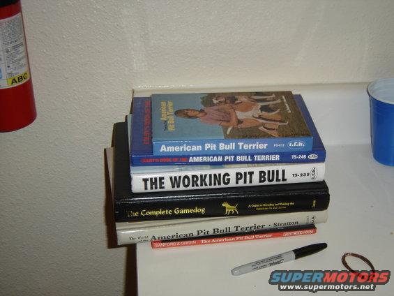 Stack of books on a shelf, including titles about pit bulls and dogs, next to a fire extinguisher and a blue cup