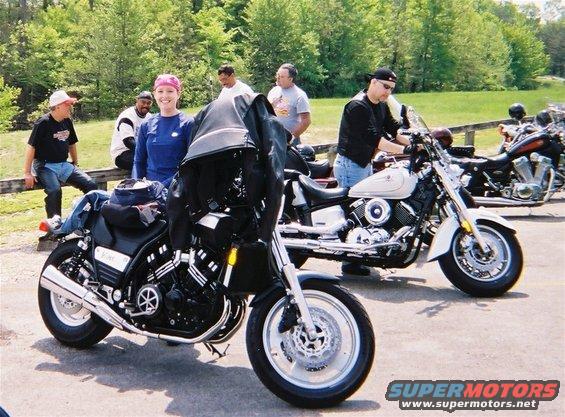 Two motorcycles parked on pavement with people standing and sitting nearby