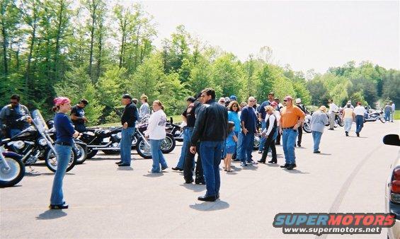 Group of people gathered around motorcycles in a parking lot with trees in the background