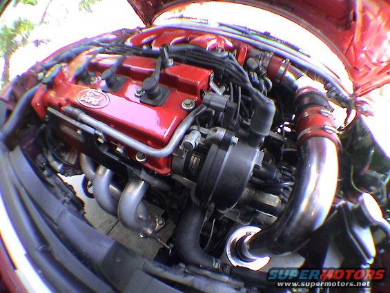 Red engine with silver exhaust headers and black hoses. Ford logo visible on valve cover