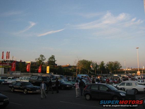 Parking lot scene with many cars and people gathered under a blue sky with wispy clouds