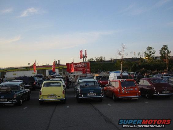 Parking lot scene with several vintage cars, including a yellow car and an orange station wagon, under a blue sky