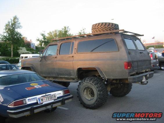 Lifted brown SUV with off-road tires and a spare tire on the roof rack parked next to a blue car