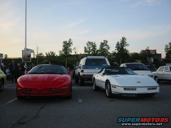 Red sports car parked in a lot, front view. Other cars and people are visible