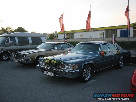 Blue Cadillac sedan with flowers on the hood and American flags on the roof, parked next to a gold sedan and a van