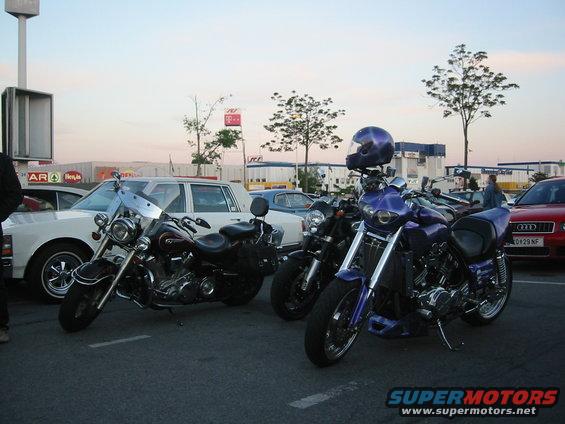 Three motorcycles parked in a lot with cars and trees