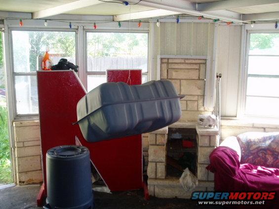 Gray plastic fuel tank suspended indoors, next to a red structure and a blue trash can