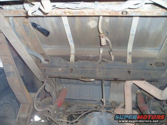 Underside of a vehicle showing rusty frame, leaf springs, red shock absorbers, and fuel lines