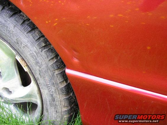 Red car fender with a dent, showing a tire and grass. Supermotors.net logo visible