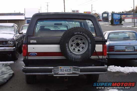 Rear view of a 1989 Ford Bronco with a spare tire mounted on the back. Another car is parked to the right