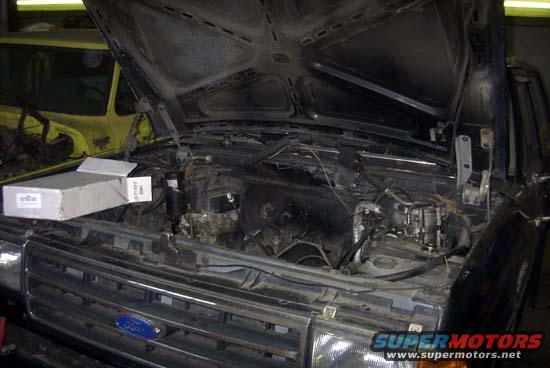 1989 Ford Bronco engine bay with hood open, showing engine components and a box on the fender. Another vehicle is visible in the background