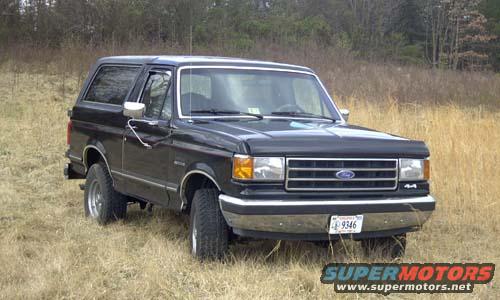 Black Ford Bronco parked in dry grass, front 3/4 view. Chrome grille, bumper, and mirrors. License plate visible