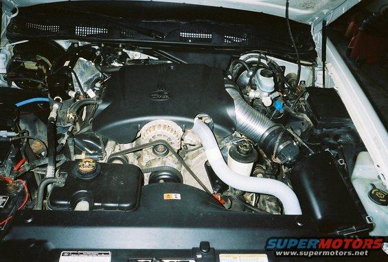 Engine bay with V8 engine components, hoses, and belts visible. Some text on labels and a watermark at the bottom