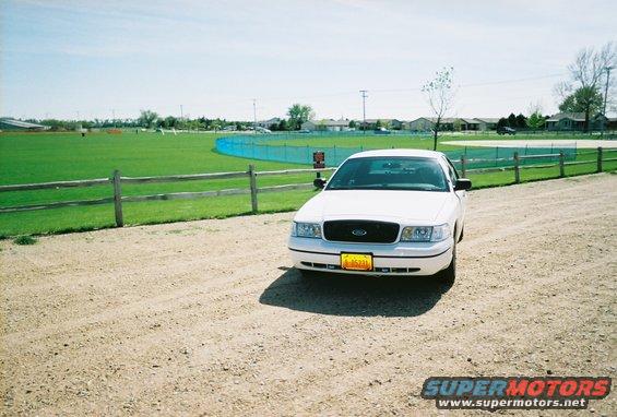 White 2000 Ford Crown Victoria parked on a dirt road near a sports field
