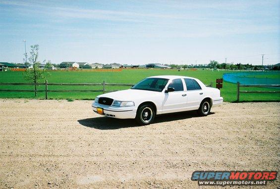 White 2000 Ford Crown Victoria parked on gravel lot with grassy field and houses in background