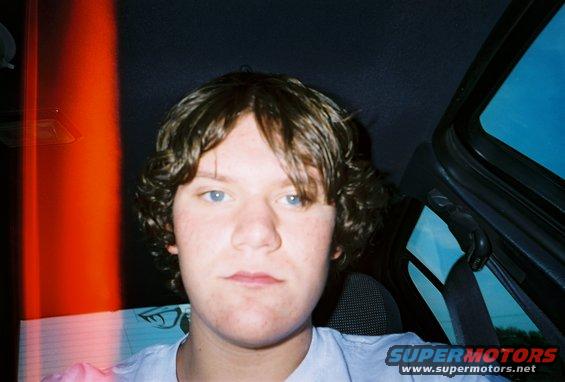 Person with curly hair and blue eyes inside a car, looking at the camera. Red light streaks on the left