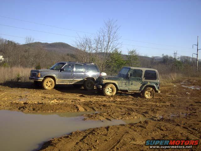 SUV and Jeep stuck in mud pit, trees and hills in background