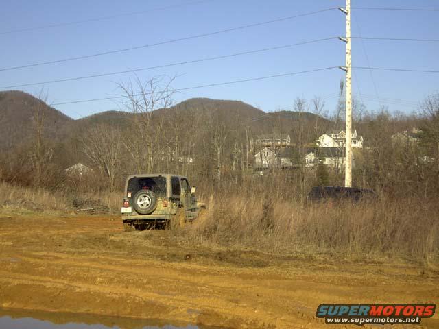 Jeep Wrangler stuck in mud with spare tire on back. Houses and trees on a hill in background