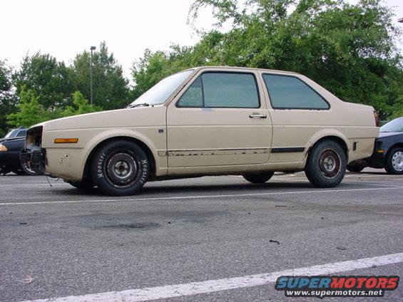 Tan two-door sedan with racing tires and missing front bumper. Appears to be a 1984 Volkswagen Golf/Jetta